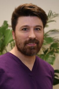 Close-up portrait of a man with brown hair and a beard wearing purple medical scrubs, with green plants in the blurred background.